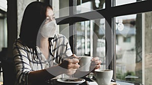 A woman wearing a medical mask sits in a coffee shop looking out the window with sad eyes