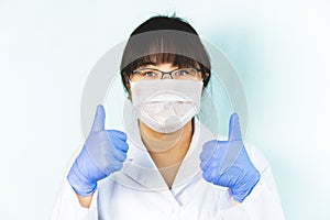 The woman is wearing a medical mask in a white uniform, protective blue gloves with a raised finger, everything will be fine