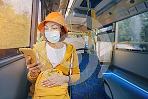 Woman wearing a medical protective mask rides a public transport bus and surfing in the Internet or application in her