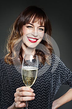 Woman wearing night party dress with a glass of champagne on dark background. Lady with long curly hair celebrating