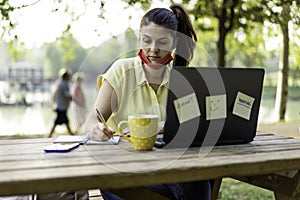Woman wearing open protective face mask using laptop outdoors - Female solopreneur taking notes while working in alternative