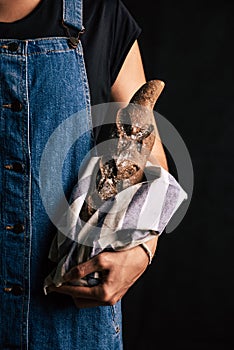 A woman wearing overalls holding a handmade rye bread wrapped with a cloth on a black background