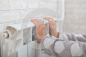 A woman wearing in pajamas warms cold hands on the heating radiator