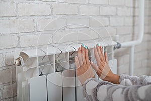 A woman wearing in pajamas warms cold hands on the heating radiator