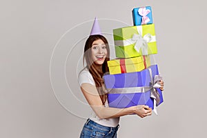 Woman wearing party cone, holding mount of gift boxes, enjoying many best presents, celebrating.