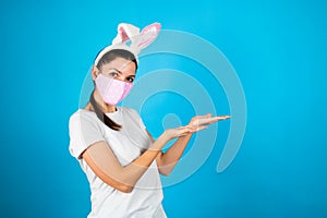 Woman wearing pink medical mask and rabbit ears  raises palms as if showing something  gestures over blank space. Coronavirus