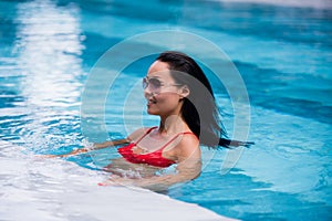 Woman wearing red swimsuit and sunglasses sitting in swimming pool, touching wet hair