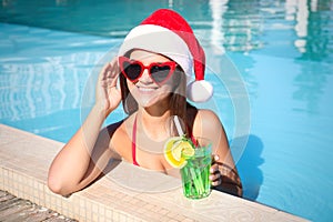 Young woman wearing Santa Claus hat with refreshing drink in swimming pool. Christmas vacation