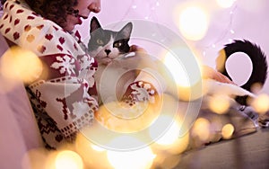 woman wearing Santa hat and Christmas sweater and adorable pet cat at home. festive decor with lights blurred