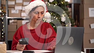 Woman wearing Santa hat working in a dropshipping office or small warehouse, preparing a parcel to deliver to the