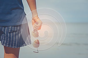 Woman wearing short pants and t-shirt standing on sand beach and holding sunglasses in hers hand.