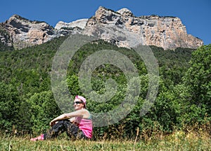 woman wearing sporty clothes, sunglasses and cap resting in an idyllic place in front of a mountain. The hiker is sitting on the