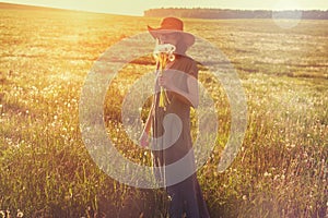 Woman wearing straw hat and long linen dress enjoying nature, holding bouquet of fresh beautiful dandelions, hot summer sunny day