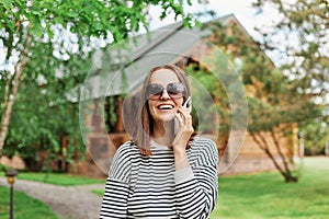 Woman wearing striped shirt and sunglasses talking on smartphone standing in park with wooden house on background laughing