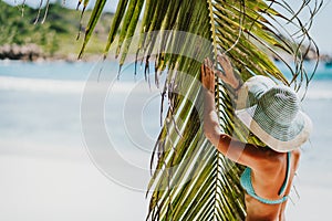 A woman wearing sun hat near coconut palm tree leaves on a tropical Anse Cocos sandy beach. La Digue, Seychelles