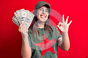 Woman wearing t-shirt with red star communist symbol holding bunch of dollars banknotes doing ok sign with fingers, smiling