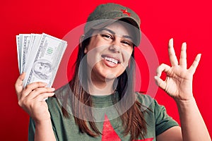 Woman wearing t-shirt with red star communist symbol holding bunch of dollars banknotes doing ok sign with fingers, smiling