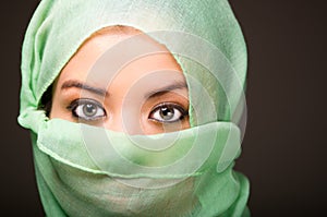 Woman wearing turquoise scarf covering face only revealing beautiful green eyes, grey dark background