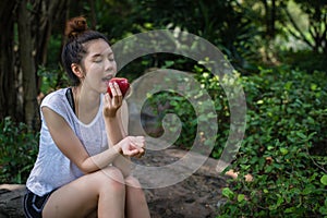 Woman is wearing white shirt and sitiing on the stone at the park and eating red apple