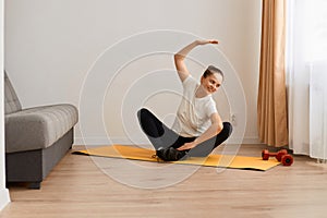 Woman wearing white t shirt and black leggins doing sport exercises at home, sitting in lotus position on mat practicing yoga,