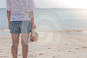 Woman wearing white t-shirt, she standing on sand beach and holding weave hat in hand, she looking at the sea.