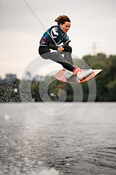 Woman in wetsuit holding rope and masterfully jumping over water on wakeboard