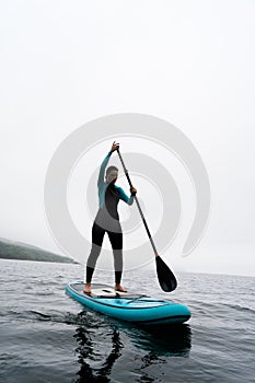 A woman in a wetsuit paddle a paddleboard with an oar on the sea waves in cloudy weather.