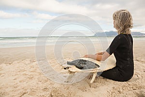 Woman in wetsuit sitting with surfboard against cloudy sky