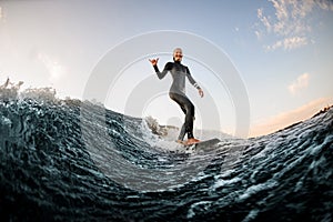 woman in wetsuit wakesurfing on the board and riding down the river wave and show hand gesture