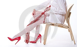 A woman with a whip and beautiful legs in white stockings and red shoes sits on a chair on a white