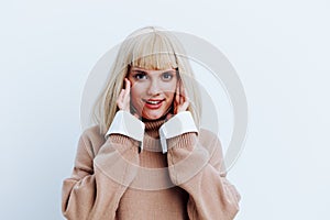 a woman on a white background in a beige sweater with beautiful blond hair looks joyfully directly covering her ears
