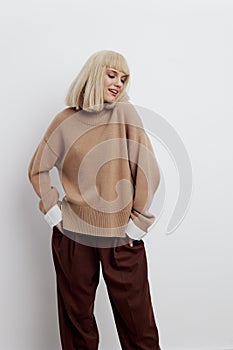 a woman on a white background in a beige sweater and brown trousers with beautiful blond hair stands and looks to the