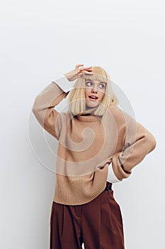 a woman on a white background in a beige sweater and brown trousers with beautiful blonde hair stands with her hand on