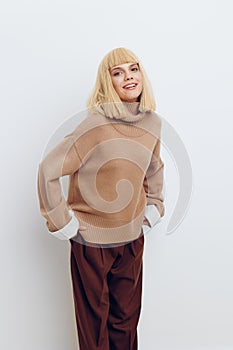 a woman on a white background in a beige sweater and brown trousers with fine blond hair stands leaning over with her