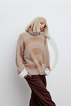 a woman on a white background in a beige sweater and brown trousers with fine blond hair stands and smiles with her