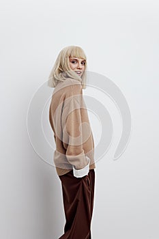 a woman on a white background in a beige sweater and brown trousers with fine blonde hair stands sideways and smiles