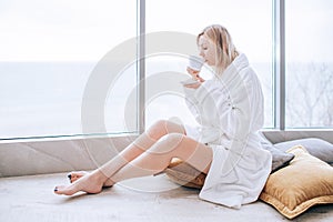 Woman in a white bathrobe enjoying sea view next to big window with cup of coffee. Panoramic window. Floor covering