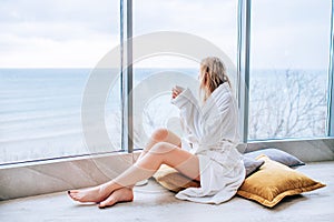 Woman in a white bathrobe enjoying sea view next to big window with cup of coffee. Panoramic window. Floor covering