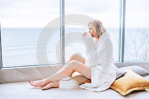 Woman in a white bathrobe enjoying sea view next to big window with cup of coffee. Panoramic window. Floor covering, Vacation in a