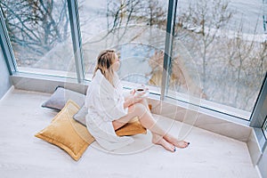 Woman in a white bathrobe enjoying sea view next to big window with cup of coffee. Panoramic window. Floor covering, Vacation in a