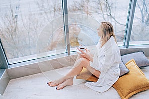 Woman in a white bathrobe enjoying sea view next to big window with cup of coffee. Panoramic window. Floor covering, Vacation