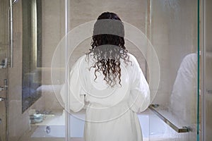 Woman in white bathrobe enters bathroom with glass