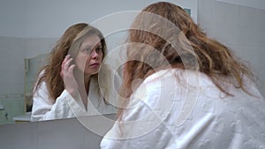 A woman in a white bathrobe looks carefully at her face in the bathroom wall mirror. Shooting from behind the shoulder.