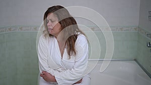 A woman in a white bathrobe, sitting on the edge of the bathtub, has a severe belly ache. A woman sits in the bathroom