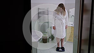 A woman in a white bathrobe stands on a bathroom scale. A woman is hysterical about her weight and steps off the