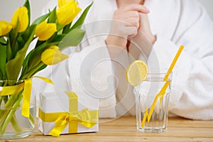 Woman in a white Bathrobe. On the table yellow tulips, gift and