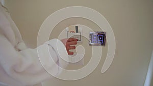 Woman in a white coat inserting electronic key card in power switch holder on wall in the hotel. Then push the button