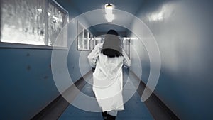 Woman in white coat running down a dark corridor, view from back.