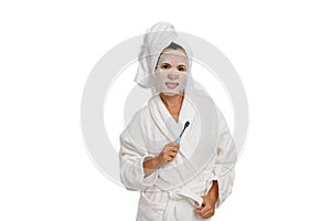 A woman in a white coat with a towel on her head, a toothbrush in her hands and a sheet mask on her face.