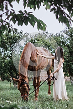 Woman in white dress bonding with tall brown horse under the green trees